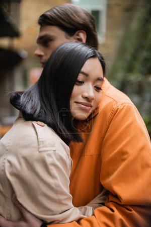 Young asian woman in trench coat hugging blurred boyfriend on terrace of cafe 
