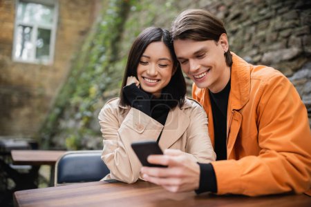 Sonriente pareja interracial usando smartphone en cafetería al aire libre 