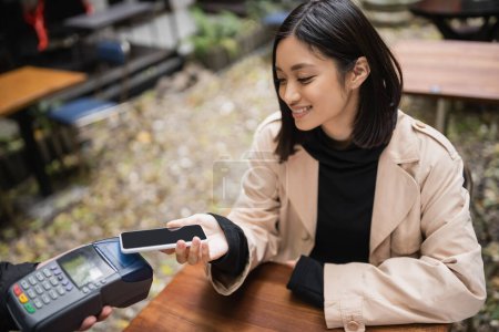 Souriant asiatique femme en trench coat payer avec smartphone près serveur tenant terminal de paiement sur terrasse café 