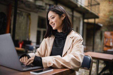 Fröhliche asiatische Bloggerin im Trenchcoat mit Laptop auf Caféterrasse 