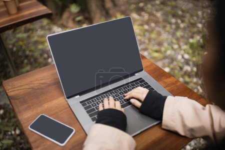 Freelancer desenfocado usando laptop con pantalla en blanco cerca de smartphone en la terraza de la cafetería 