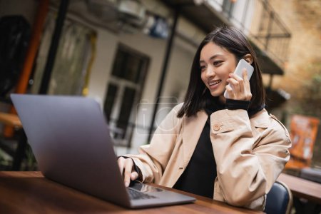 Positive asian freelancer talking on smartphone near blurred laptop in outdoor cafe 