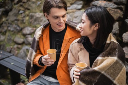 Foto de Sonriente joven sosteniendo café para ir y mirando asiática novia en manta en terraza de café - Imagen libre de derechos
