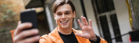 Téléchargez les photos : Jeune homme positif ayant appel vidéo sur smartphone dans un café extérieur, bannière - en image libre de droit