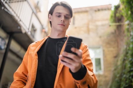 Téléchargez les photos : Vue à angle bas du jeune homme en veste à l'aide d'un smartphone flou sur la terrasse du café - en image libre de droit