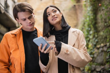 Low angle view of asian woman pointing at smartphone near boyfriend outdoors 