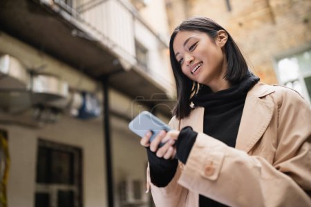 Foto de Bajo ángulo vista de sonriente mujer asiática en gabardina usando teléfono inteligente al aire libre - Imagen libre de derechos