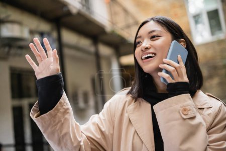 Lächelnde Asiatin, die auf dem Smartphone spricht und draußen mit der Hand winkt 