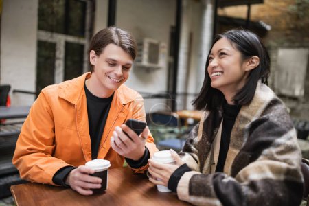 Foto de Hombre positivo usando teléfono inteligente cerca de novia asiática en manta con café en la terraza de la cafetería - Imagen libre de derechos