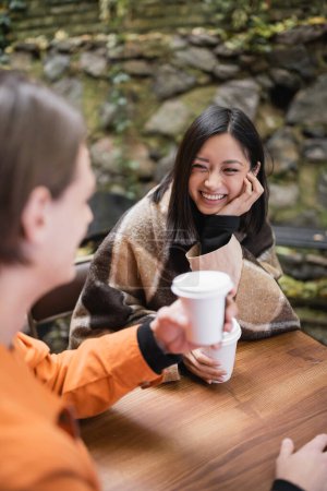 Cheerful asian woman in blanket holding coffee to go near blurred boyfriend in outdoor cafe 