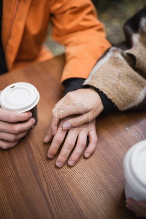 Cropped view of woman in blanket touching hand of boyfriend near coffee to go in outdoor cafe 