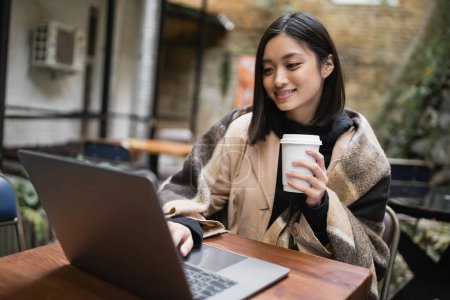 Smiling asian woman in blanket holding coffee to go and using laptop on terrace of cafe 