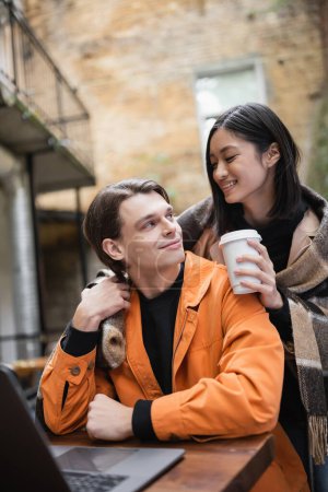 Positivo asiático mujer sosteniendo papel taza cerca novio y borrosa portátil en terraza de café 