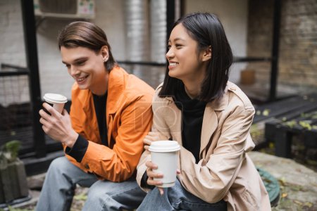 Téléchargez les photos : Gai asiatique femme en trench coat tenant café pour aller près copain sur café terrasse - en image libre de droit