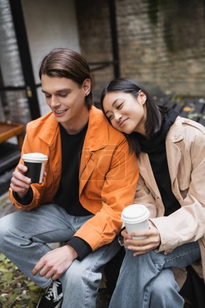 Smiling man looking at coffee to go near pleased asian girlfriend in trench coat in outdoor cafe 