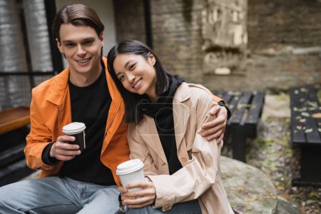 Foto de Positivo hombre sosteniendo papel taza y abrazando asiático novia en gabardina en al aire libre café - Imagen libre de derechos
