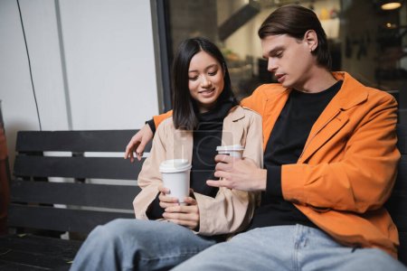 Foto de Joven pareja interracial sosteniendo café para ir mientras está sentado en el banco en la cafetería al aire libre - Imagen libre de derechos