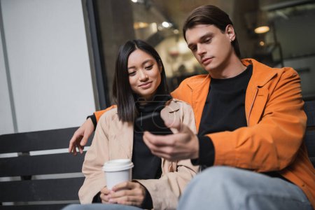 Foto de Joven pareja multiétnica con café para llevar y smartphone sentado en el banco en la terraza de la cafetería - Imagen libre de derechos