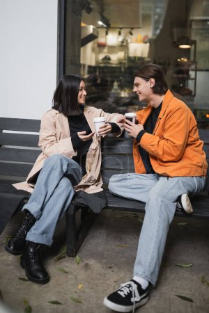 Stylish interracial couple holding coffee to go while talking on bench near cafe 