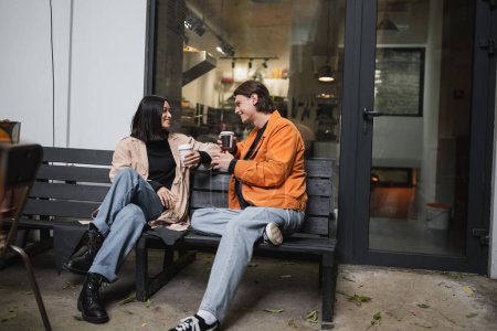 Young multiethnic couple with coffee to go talking on bench on cafe terrace 
