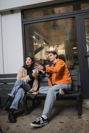 Junger Mann zeigt mit dem Finger, während er Kaffee hält, um in die Nähe einer asiatischen Freundin auf einer Bank in der Nähe eines Cafés zu gehen 