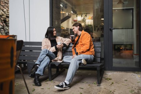 Trendy multiethnic couple talking while holding takeaway cups on bench near cafe 