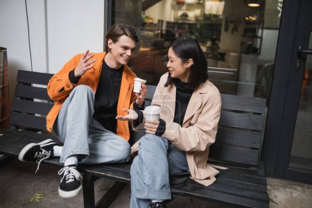 Young multiethnic couple holding paper cups while talking on bench near cafe 