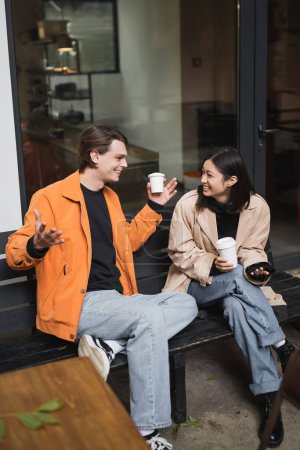 Cheerful asian woman talking to boyfriend with coffee to go on bench near cafe 