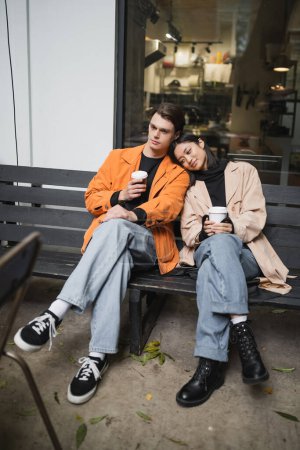 Foto de Joven mujer asiática en gabardina sosteniendo café para ir cerca de novio en banco cerca de cafetería - Imagen libre de derechos