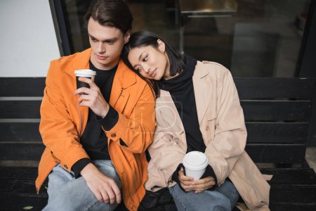 Junger Mann hält Kaffee für asiatische Freundin im Trenchcoat auf Bank in der Nähe von Café 