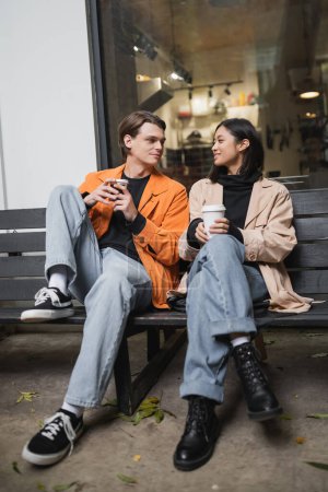 Positive multiethnic couple talking while holding coffee on bench on terrace of cafe 