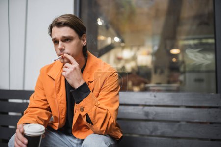 Stylish man smoking cigarette and holding coffee to go near cafe 