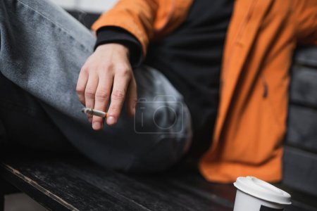 Cropped view of blurred man holding cigarette near coffee to go on bench outdoors 