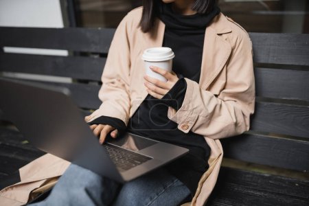 Foto de Vista recortada de la mujer joven en gabardina sosteniendo la taza de papel y utilizando el ordenador portátil en el banco al aire libre - Imagen libre de derechos