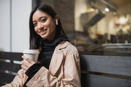 Smiling asian woman in trench coat holding coffee to go and looking at camera on bench near cafe 