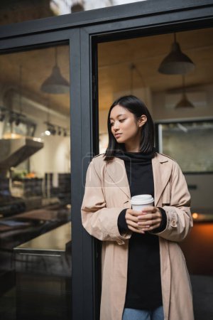 Stilvolle asiatische Frau im Trenchcoat hält Kaffee in der Nähe der Tür eines Cafés 