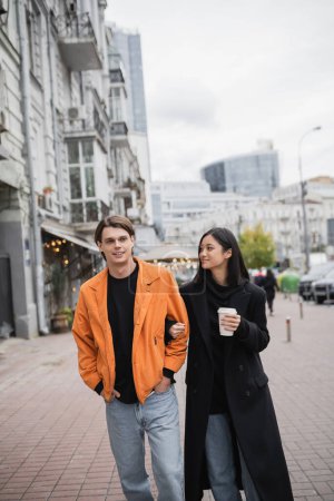 Smiling asian woman holding coffee to go and looking at boyfriend on urban street 