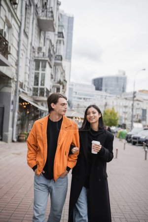 Stylish man looking at cheerful asian girlfriend with coffee to go on urban street 