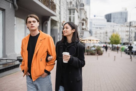Stylish young interracial couple with coffee to go walking on urban street 