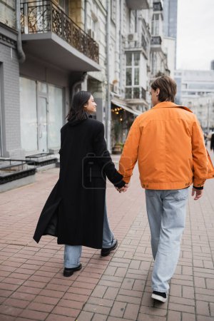 Side view of smiling interracial couple holding hands while walking on urban street 