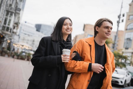 Foto de Positivo asiático mujer holding takeaway café mientras caminando cerca novio en urbano calle - Imagen libre de derechos