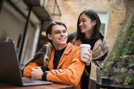 Junge asiatische Frau in Decke hält Kaffee to go und umarmt Freund in der Nähe von Laptop auf der Terrasse des Cafés 