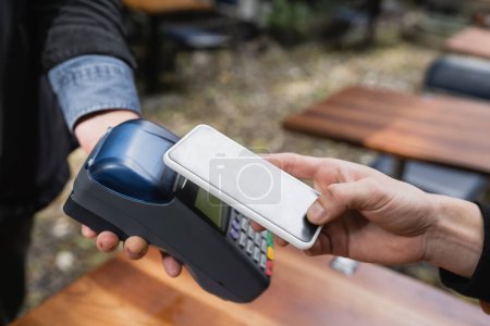 Ausgeschnittene Ansicht von Mann, der mit Handy in der Nähe von Kellnerin mit Kreditkartenlesegerät in Outdoor-Café bezahlt 