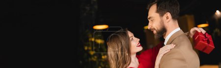 Foto de Vista lateral de mujer alegre sosteniendo presente y abrazando novio barbudo en día de San Valentín, bandera - Imagen libre de derechos