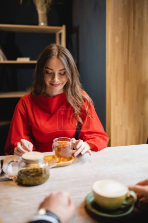 fröhliche junge Frau im roten Kleid blickt auf Glasschale mit grünem Tee in der Nähe ihres Freundes  