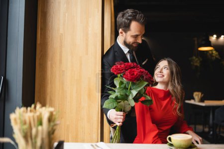 Foto de Hombre barbudo de traje con ramo de rosas rojas cerca de la novia sonriente en el día de San Valentín - Imagen libre de derechos
