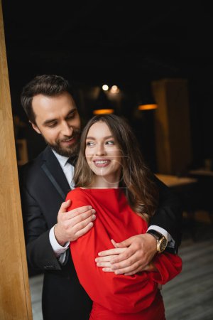 bärtiger Mann im Anzug umarmt fröhliche Frau im roten Kleid am Valentinstag 