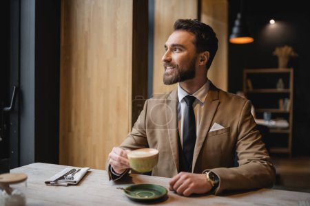 happy bearded man in suit holding cup of cappuccino in cafe 