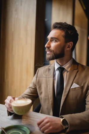 bearded man in suit holding cup of cappuccino and looking away in cafe 