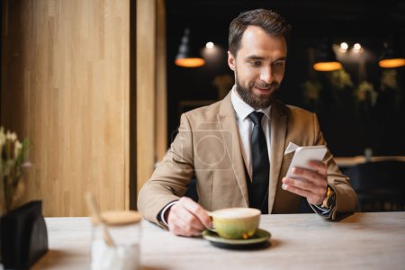 Photo for Cheerful man with beard using smartphone while holding cup of cappuccino in cafe - Royalty Free Image